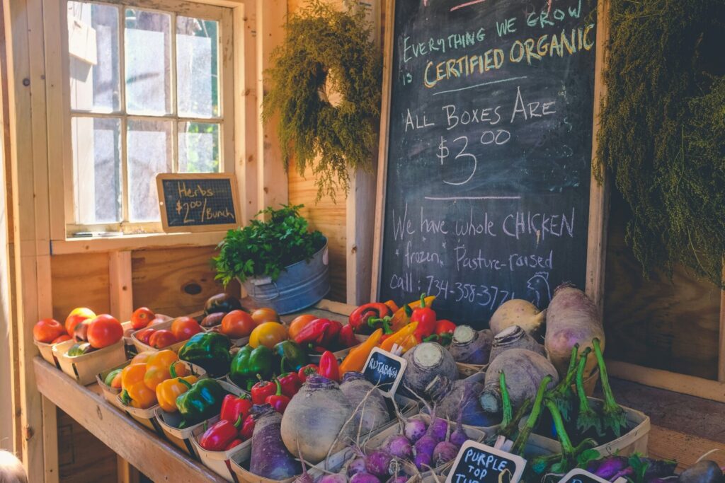 A market stall with various vegetables, including bell peppers, beets, and radishes, displayed with price tags. A chalkboard announces certified organic produce at $3 per box. Support local businesses like Sammamish Carpet Cleaning and community partners for all your needs.