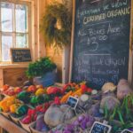 A market stall with various vegetables, including bell peppers, beets, and radishes, displayed with price tags. A chalkboard announces certified organic produce at $3 per box. Support local businesses like Sammamish Carpet Cleaning and community partners for all your needs.