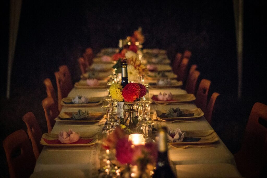 A long dining table set for a meal with plates, glasses, napkins, candles, and flower centerpieces, surrounded by empty chairs in dim lighting.