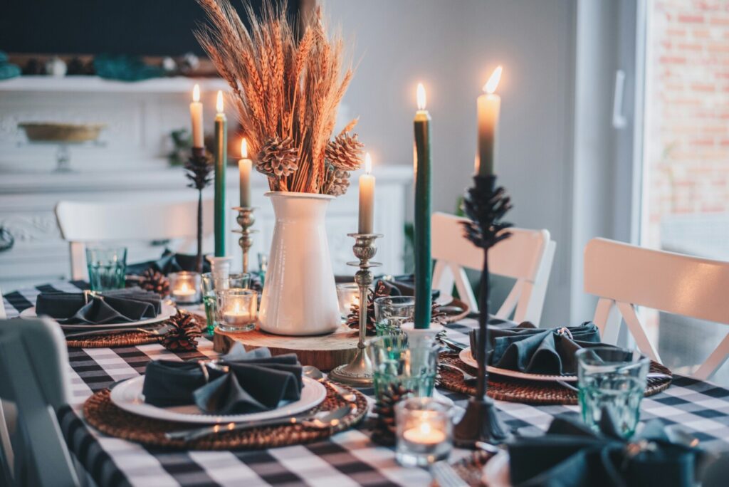 A dining table set for a meal features checkered tablecloth, black napkins, glassware, lit candles, and a central arrangement of dried wheat in a white vase.