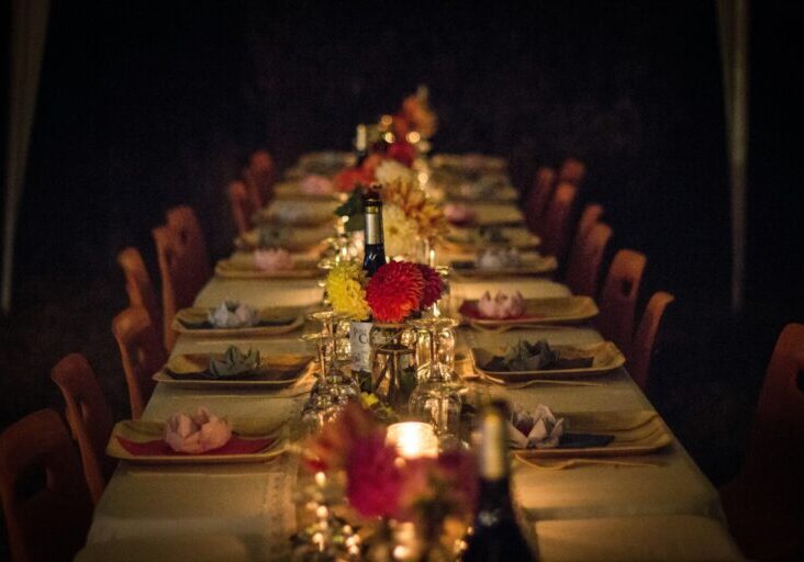A long dining table set for a meal with plates, glasses, napkins, candles, and flower centerpieces, surrounded by empty chairs in dim lighting.