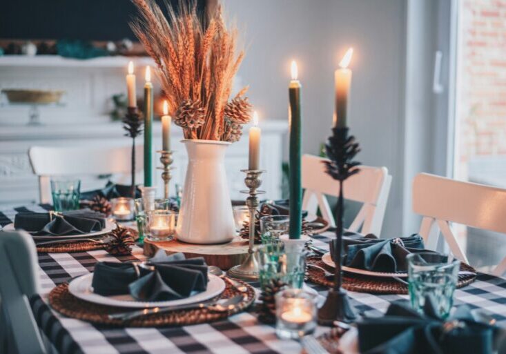 A dining table set for a meal features checkered tablecloth, black napkins, glassware, lit candles, and a central arrangement of dried wheat in a white vase.